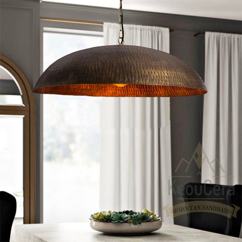May include: A large, hammered, brass pendant light fixture hangs from a chain in a modern dining room. The light fixture is round and has a warm, golden glow. The light fixture is suspended over a table with a white tablecloth and a bowl of succulents.