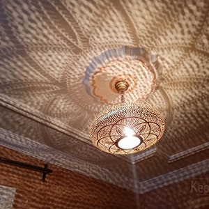 May include: A gold Moroccan pendant light fixture with intricate geometric patterns casts a beautiful shadow on the ceiling. The light fixture is hanging from a chain and has a white light bulb inside.