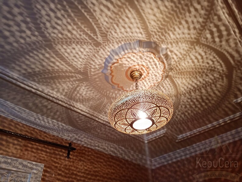 May include: A gold Moroccan pendant light fixture with intricate geometric patterns casts a beautiful shadow on the ceiling. The light fixture is hanging from a chain and has a white light bulb inside.