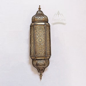 May include: A gold metal Moroccan lantern with intricate cut-out designs. The lantern has a domed top and a rectangular body. It is a traditional style lantern that would be perfect for adding a touch of Moroccan style to any home.