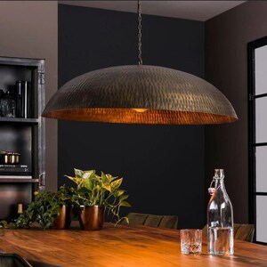 May include: A large, round, hammered metal pendant light fixture hangs over a wooden table. The light fixture is a warm, brassy color and has a textured surface. The table is surrounded by plants in pots.