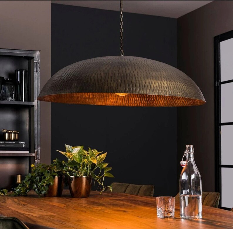 May include: A large, round, hammered metal pendant light fixture hangs over a wooden table. The light fixture is a warm, brassy color and has a textured surface. The table is surrounded by plants in pots.