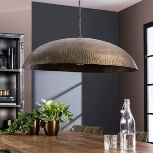 May include: A large, hammered brass pendant light fixture hangs over a wooden table with plants and a glass of water. The light fixture has a wide, dome-shaped shade.