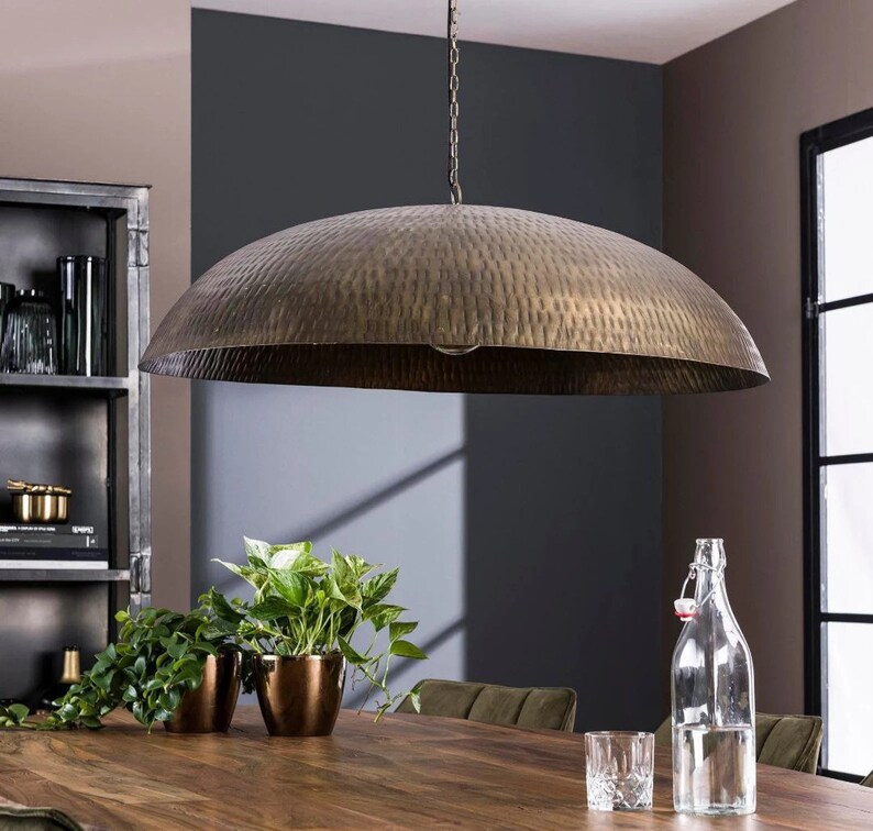 May include: A large, hammered brass pendant light fixture hangs over a wooden table with plants and a glass of water. The light fixture has a wide, dome-shaped shade.