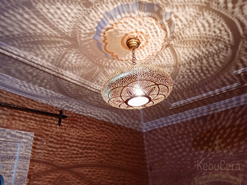 May include: A gold-toned Moroccan pendant light fixture with intricate cut-out designs hangs from a chain in a room with a white ceiling and brown walls. The light casts a patterned shadow on the walls.