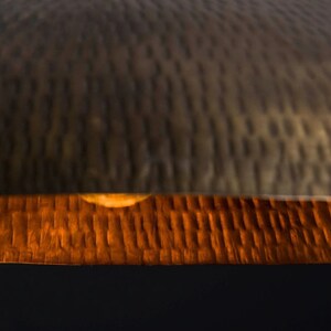 May include: A close-up of a hammered metal lampshade with a warm, orange glow emanating from the bottom. The lampshade is made of a textured metal with a slightly distressed finish.