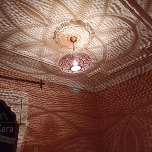 May include: A Moroccan-style pendant light fixture with a round, openwork design. The light casts intricate patterns on the ceiling and walls, creating a warm and inviting atmosphere. The fixture is made of metal and has a brass finish. The light is hanging from a chain and is suspended from a ceiling medallion.