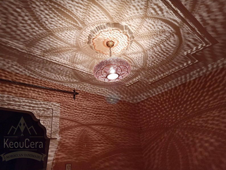 May include: A Moroccan-style pendant light fixture with a round, openwork design. The light casts intricate patterns on the ceiling and walls, creating a warm and inviting atmosphere. The fixture is made of metal and has a brass finish. The light is hanging from a chain and is suspended from a ceiling medallion.