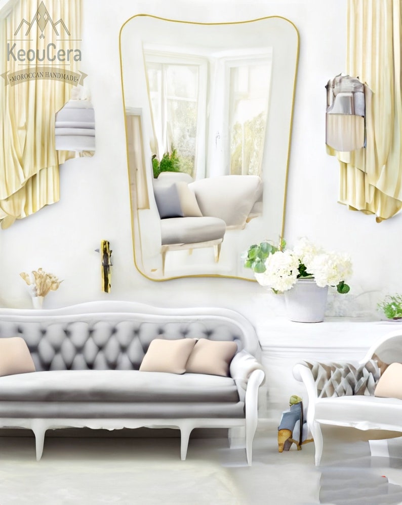 May include: A white, tufted sofa with a curved back and four legs. The sofa has two light pink pillows and is in a room with a large, gold-framed mirror and white walls.