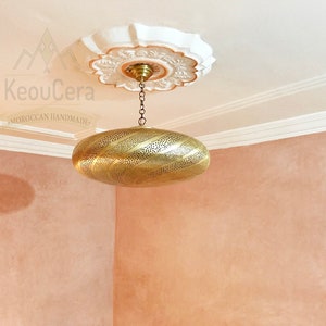 May include: A gold-toned, oval-shaped pendant light fixture with intricate cut-out designs hangs from a chain against a light pink wall. The light fixture is suspended from a white ceiling medallion with a floral design. The text "KeouCera Moroccan Handmade" is visible in the image.