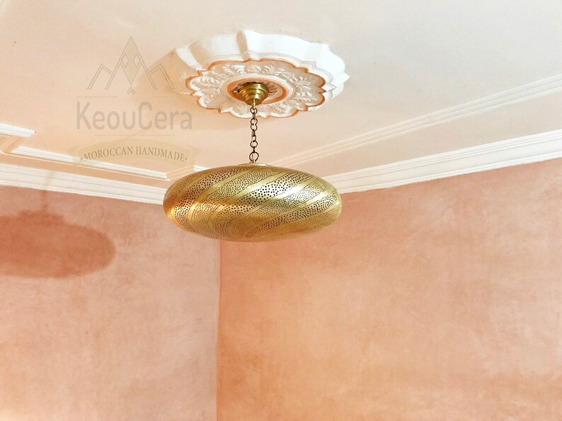 May include: A gold-toned, oval-shaped pendant light fixture with intricate cut-out designs hangs from a chain against a light pink wall. The light fixture is suspended from a white ceiling medallion with a floral design. The text "KeouCera Moroccan Handmade" is visible in the image.