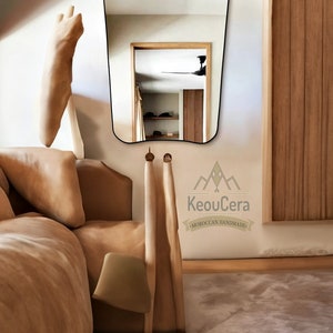 May include: A tan leather bed with a white pillow and a tan throw blanket. A wooden chair with a tan fabric seat is next to the bed. A large, wavy-shaped mirror hangs on the wall behind the bed. The mirror reflects a room with a ceiling fan and a wooden door. The text "KeouCera - MOROCCAN HANDMADE" is printed on the wall below the mirror.
