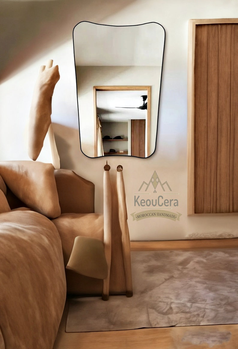 May include: A tan leather bed with a white pillow and a tan throw blanket. A wooden chair with a tan fabric seat is next to the bed. A large, wavy-shaped mirror hangs on the wall behind the bed. The mirror reflects a room with a ceiling fan and a wooden door. The text "KeouCera - MOROCCAN HANDMADE" is printed on the wall below the mirror.