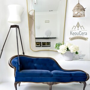 May include: A blue velvet chaise lounge with wooden legs and a curved back. The chaise lounge is in front of a white wall with a gold-framed mirror and a white lamp.