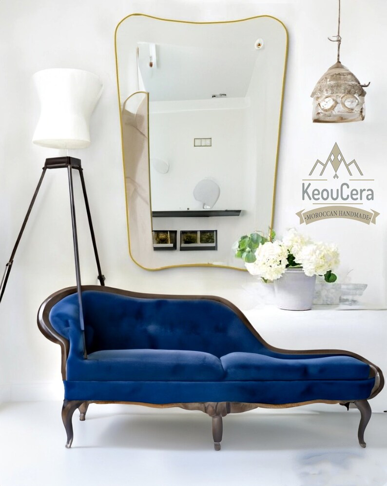 May include: A blue velvet chaise lounge with wooden legs and a curved back. The chaise lounge is in front of a white wall with a gold-framed mirror and a white lamp.
