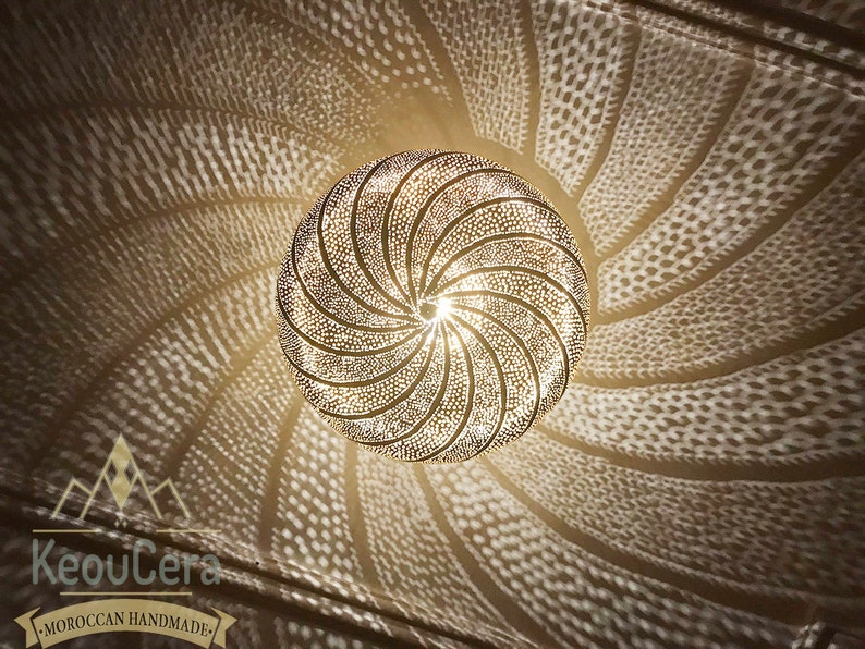 May include: A gold-toned Moroccan pendant light fixture with a circular, geometric design. The light fixture casts intricate shadows on the ceiling.