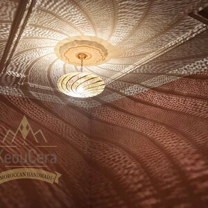 May include: A gold Moroccan pendant light fixture with a geometric design casts intricate shadows on a white ceiling. The light fixture is hanging from a chain and has a round shape. The shadows are a mix of light and dark, creating a beautiful pattern.