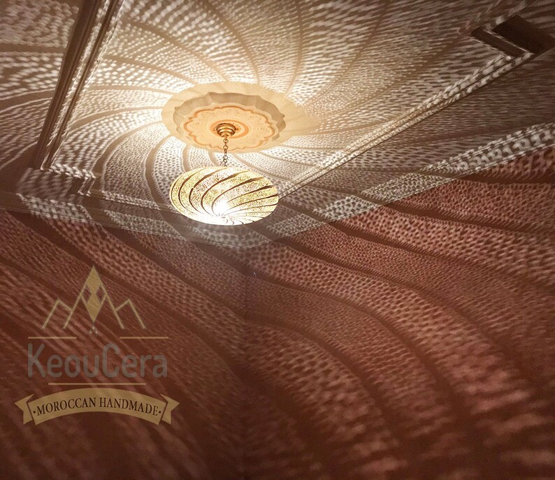 May include: A gold Moroccan pendant light fixture with a geometric design casts intricate shadows on a white ceiling. The light fixture is hanging from a chain and has a round shape. The shadows are a mix of light and dark, creating a beautiful pattern.