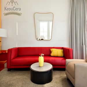 May include: A red sofa with a yellow pillow sits in a living room with a white wall and a gold-framed mirror. A yellow vase with a flower sits on a black and white coffee table in front of the sofa. A beige armchair sits to the right of the sofa.