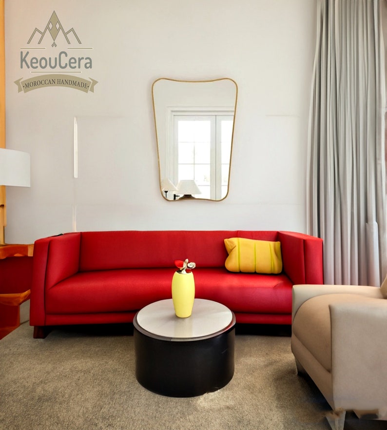 May include: A red sofa with a yellow pillow sits in a living room with a white wall and a gold-framed mirror. A yellow vase with a flower sits on a black and white coffee table in front of the sofa. A beige armchair sits to the right of the sofa.