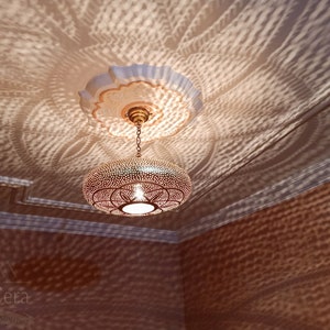 May include: A round, brass pendant light with a geometric pattern hangs from a chain. The light casts intricate shadows on the ceiling.