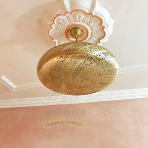 May include: A gold-toned, round, Moroccan-style pendant light fixture with intricate cut-out designs. The light fixture is hanging from a white ceiling with ornate molding. The text "KeouCera -MOROCCAN HANDMADE- " is visible below the light fixture.