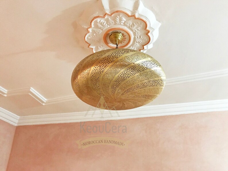 May include: A gold-toned, round, Moroccan-style pendant light fixture with intricate cut-out designs. The light fixture is hanging from a white ceiling with ornate molding. The text "KeouCera -MOROCCAN HANDMADE- " is visible below the light fixture.