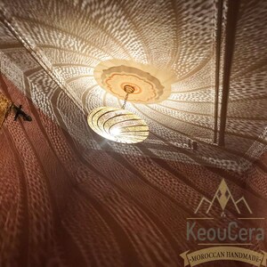 May include: A gold Moroccan pendant light fixture with a geometric design casts intricate shadows on a brown wall. The light fixture is hanging from a ceiling with a white and gold painted design. The text "KeouCera - MOROCCAN HANDMADE" is visible in the bottom right corner of the image.