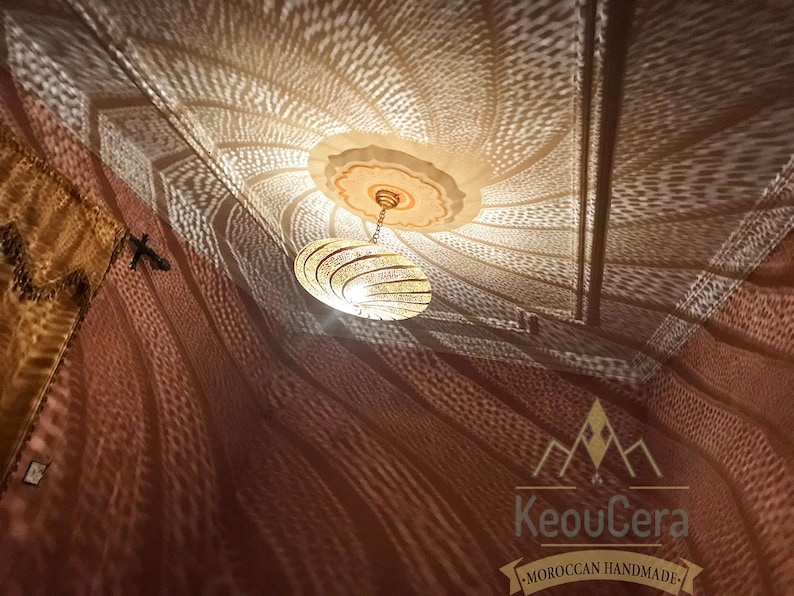 May include: A gold Moroccan pendant light fixture with a geometric design casts intricate shadows on a brown wall. The light fixture is hanging from a ceiling with a white and gold painted design. The text "KeouCera - MOROCCAN HANDMADE" is visible in the bottom right corner of the image.