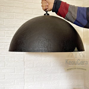 May include: A black, hammered metal dome pendant light fixture hanging from a ceiling. The light fixture has a textured surface and is suspended by a black cord.