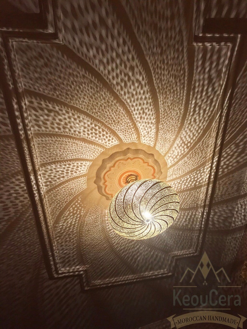 May include: A gold metal Moroccan pendant light with a geometric pattern casts a shadow on the ceiling. The light is hanging from a chain and has a round shape. The shadow is a repeating pattern of small circles.