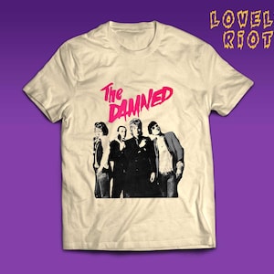 Peut inclure: T-shirt crème avec une image en noir et blanc d'un groupe et le texte rose "The Damned". Le texte "LOVELY RIOT" est en jaune en haut à droite.