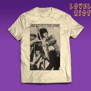 Könnte beinhalten: Cremefarbenes T-Shirt mit einem Schwarz-Weiß-Bild einer Band. Der Aufdruck beinhaltet den Text "KICK'N OUT THE JAMS" und den Markennamen "LOVELY RIOT". Das Shirt wird vor einem violetten Hintergrund präsentiert, im Rock-Stil.