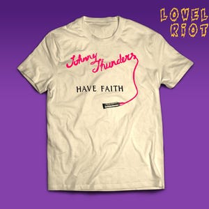 Puede incluir: Camiseta color crema con la frase "Johnny Thunders" en escritura rosa sobre "HAVE FAITH" en negro. Una línea rosa se extiende desde el texto hasta un gráfico negro y rojo. Las palabras "LOVELY RIOT" están en la esquina superior derecha.