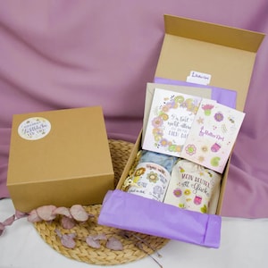 Könnte beinhalten: Eine braune Kartonbox mit einer violetten Seidenpapierauskleidung. Die Box enthält eine kleine weiße Tasche mit dem Text "Mein Beutel voll Glück" und ein kleines Glas mit dem Text "Wünsch dir was" und einer bunten Illustration eines Elefanten. Die Box enthält auch eine Karte mit dem Text "Du bist nicht allein, ich bin für dich da" und eine kleine Karte mit dem Text "Rollin' Art".