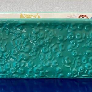 K&ouml;nnte beinhalten: Eine rechteckige t&uuml;rkisfarbene Keramik-Backform mit einem strukturierten Blumenmuster auf der Au&szlig;enseite. Die Form hat zwei kleine Griffe und einen wei&szlig;en Innenraum mit floralen Akzenten. Dieser K&uuml;chenartikel eignet sich zum Backen und Servieren.