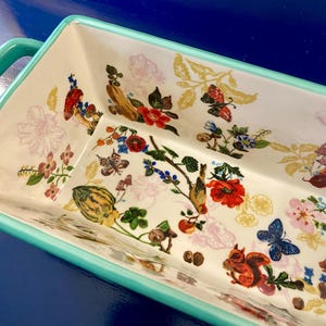 K&ouml;nnte beinhalten: Rechteckige Keramik-Backform mit wei&szlig;em Innenraum und farbenfrohem Blumen- und Tiermotiv. Das Design umfasst rote Mohnblumen, Schmetterlinge und ein Eichh&ouml;rnchen. Die Form hat einen t&uuml;rkisfarbenen Rand und Griff.