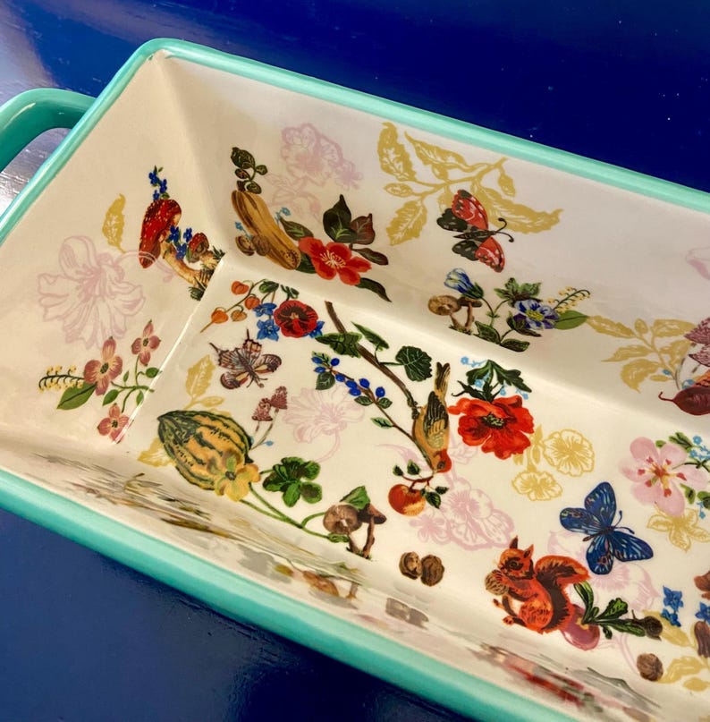 K&ouml;nnte beinhalten: Rechteckige Keramik-Backform mit wei&szlig;em Innenraum und farbenfrohem Blumen- und Tiermotiv. Das Design umfasst rote Mohnblumen, Schmetterlinge und ein Eichh&ouml;rnchen. Die Form hat einen t&uuml;rkisfarbenen Rand und Griff.