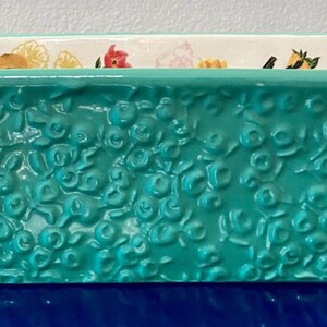 K&ouml;nnte beinhalten: Eine rechteckige t&uuml;rkisfarbene Keramik-Backform mit gepr&auml;gten floralen Details. Die Form hat zwei Griffe und ein Blumenmuster am Innenrand. Dieser K&uuml;chenartikel eignet sich zum Backen und Servieren.