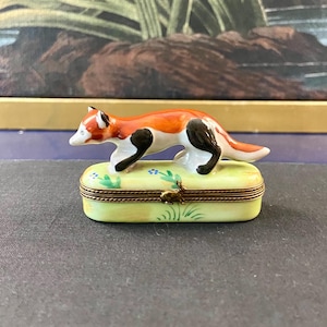 May include: A ceramic trinket box with a fox figurine on top. The fox is orange, black, and white. The box is light yellow with a floral design and a gold-colored clasp. The box is rectangular.