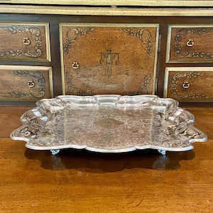 May include: A silver-colored, rectangular serving tray with ornate, scalloped edges and small feet. The tray has a detailed, embossed pattern across its surface. The background features a wooden cabinet with decorative panels and brass hardware.