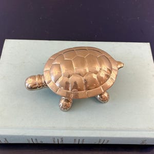 May include: A polished brass turtle figurine with a detailed, segmented shell pattern. The turtle has a smooth, reflective metallic surface, a rounded head, and four short legs, capturing a realistic shape in a compact, decorative metal form.