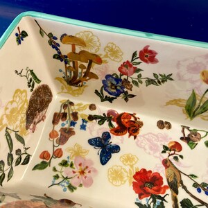 K&ouml;nnte beinhalten: Rechteckige Keramikservierschale mit wei&szlig;em Boden und hellgr&uuml;nem Rand. Die Schale ist mit einem farbenfrohen Blumen- und Tiermuster verziert, darunter Pilze, Blumen, ein Schmetterling und ein Igel.
