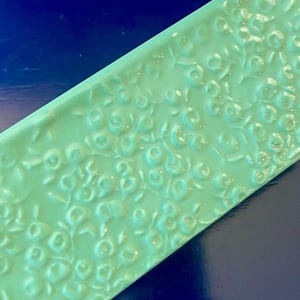 K&ouml;nnte beinhalten: Eine rechteckige, hellgr&uuml;ne Fliese mit einem erhabenen Blumenmuster. Die Oberfl&auml;che der Fliese ist gl&auml;nzend, und das gepr&auml;gte Design zeigt kleine, runde bl&uuml;tenartige Formen und zarte Bl&auml;tter. Die Fliese ist vor einem dunkelblauen Hintergrund platziert.
