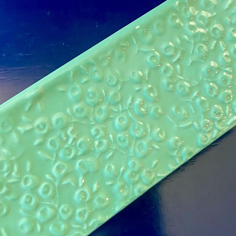 K&ouml;nnte beinhalten: Eine rechteckige, hellgr&uuml;ne Fliese mit einem erhabenen Blumenmuster. Die Oberfl&auml;che der Fliese ist gl&auml;nzend, und das gepr&auml;gte Design zeigt kleine, runde bl&uuml;tenartige Formen und zarte Bl&auml;tter. Die Fliese ist vor einem dunkelblauen Hintergrund platziert.