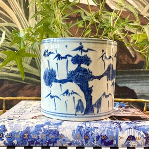 May include: A cylindrical ceramic planter featuring a white base with blue hand-painted illustrations of pine trees and mountain landscapes. The planter has a subtle crackle glaze finish and is decorated with horizontal blue bands at the top and bottom.