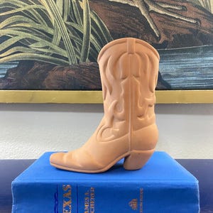 May include: A tan-colored cowboy boot figurine with decorative stitching details. The boot is displayed on a blue book with gold lettering. The background features a framed painting with a nature scene.