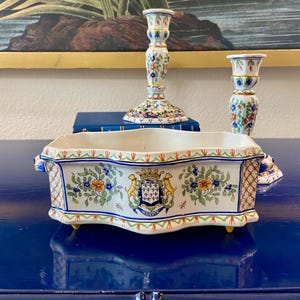 May include: A rectangular ceramic planter with a white base, adorned with floral patterns in blue, orange, and green. It features a crest design and is flanked by two matching candlesticks, all on a blue surface.