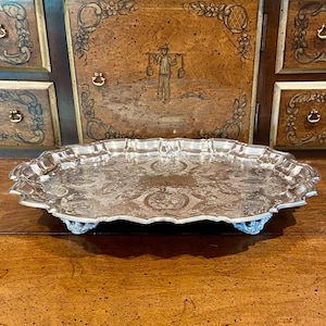 May include: A silver-colored, oval serving tray with ornate floral patterns and a scalloped edge. The tray is supported by four decorative feet. The background features a wooden cabinet with painted details.