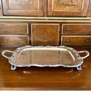 May include: A rectangular silver-toned serving tray featuring ornate, scrolled handles and decorative feet. The tray has a polished, reflective surface with an intricate, engraved floral pattern centered on the base and a raised, scalloped edge.